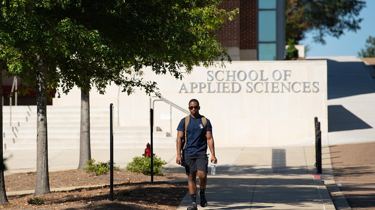 Student walks along a campus sidewalk carrying a backpack and water bottle in front of the School of Applied Sciences building at the University of Mississippi. Trees line the walkway and the school's name is visible on the building behind him.