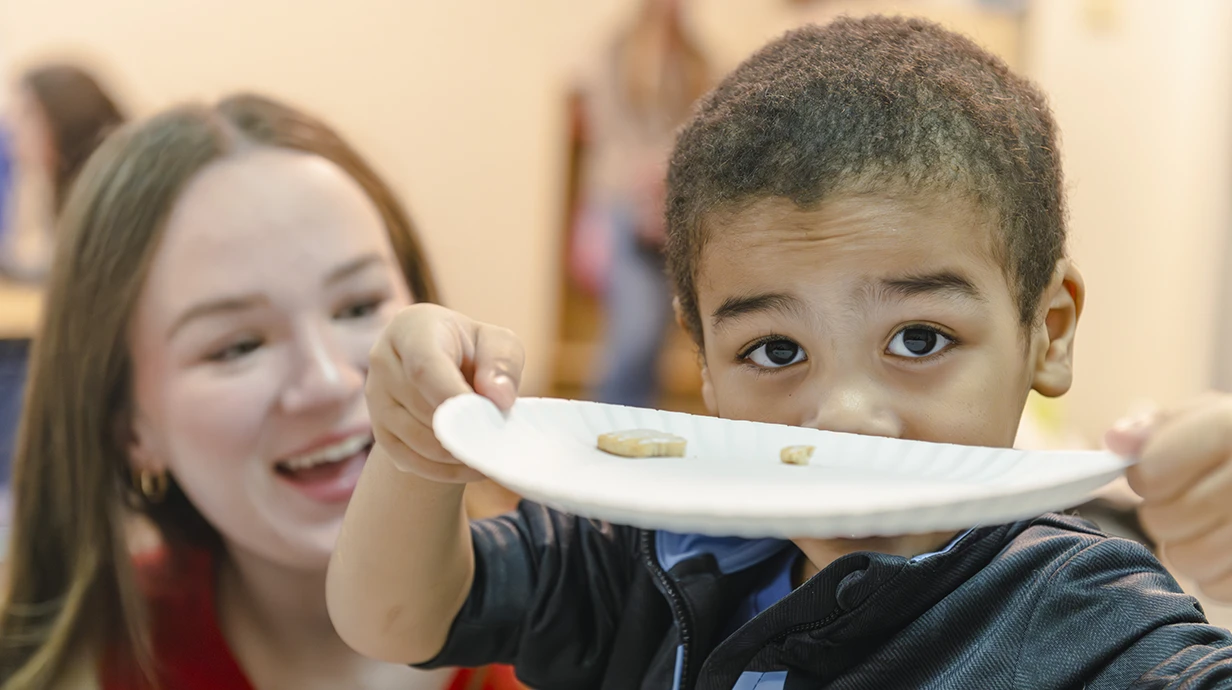 A student laughs as a child in a communications sciences and disorders therapy sessions holds up his plate with animal crackers on it to the camera in a goofy fashion