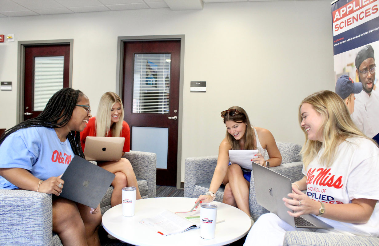 Image of four young, diverse women in the School of Applied Sciences gathered around round table looking at notebook